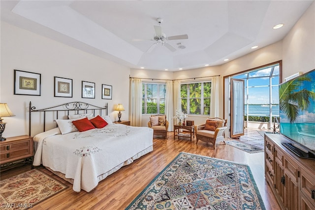 bedroom featuring ceiling fan, a raised ceiling, access to outside, a water view, and hardwood / wood-style flooring