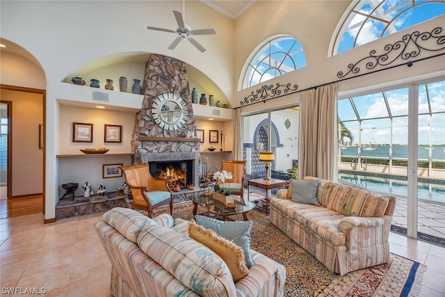 tiled living room with a towering ceiling, ceiling fan, a stone fireplace, and a wealth of natural light