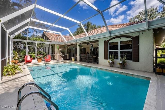 view of swimming pool featuring glass enclosure, ceiling fan, an outdoor kitchen, a grill, and a patio