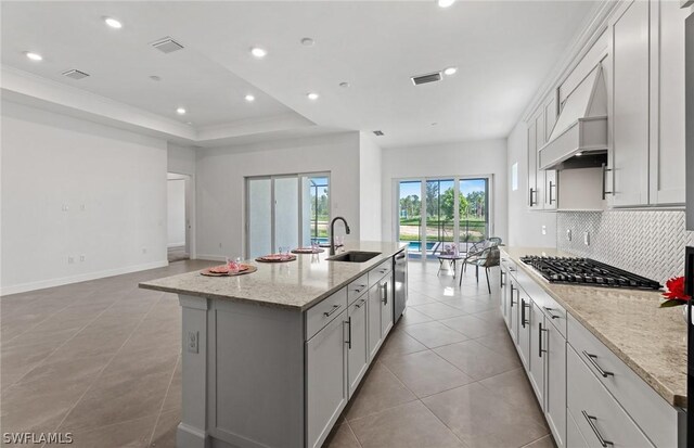 kitchen featuring light stone countertops, custom range hood, stainless steel appliances, sink, and an island with sink