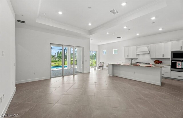 kitchen featuring custom exhaust hood, white cabinetry, an island with sink, and a tray ceiling