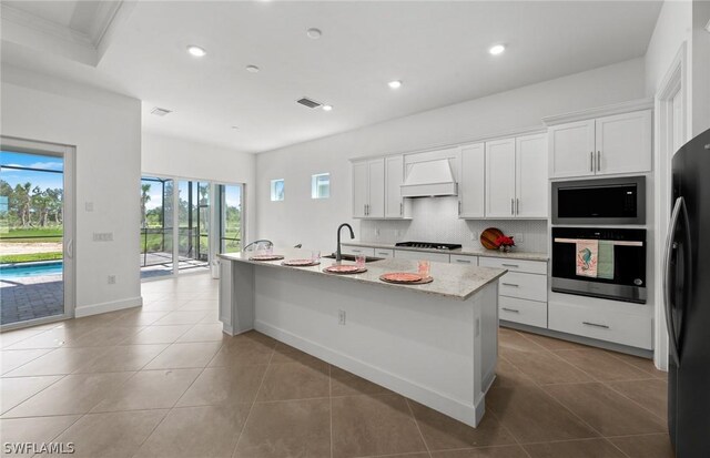 kitchen featuring black appliances, white cabinetry, an island with sink, and premium range hood