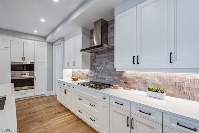 kitchen featuring stainless steel appliances, wall chimney exhaust hood, white cabinets, backsplash, and light wood-type flooring