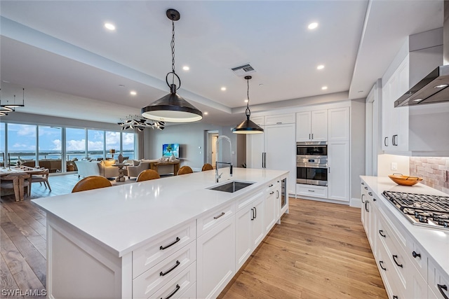 kitchen featuring an island with sink, sink, wall chimney range hood, decorative light fixtures, and white cabinetry