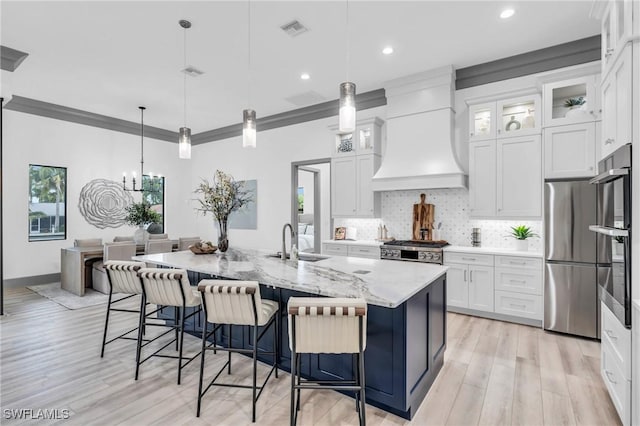 kitchen featuring a center island with sink, sink, custom exhaust hood, white cabinetry, and hanging light fixtures