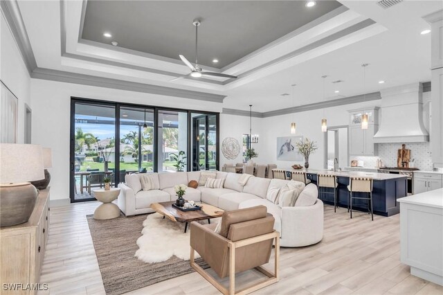 living room with light wood-type flooring, sink, ornamental molding, ceiling fan, and a raised ceiling