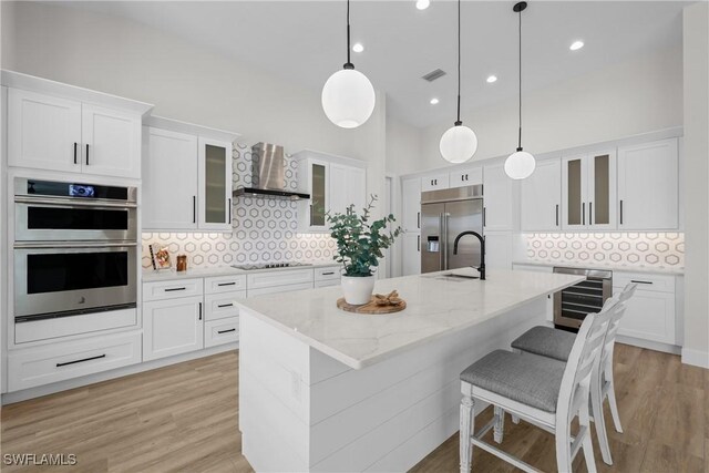 kitchen featuring backsplash, appliances with stainless steel finishes, light hardwood / wood-style floors, wall chimney range hood, and a kitchen island with sink