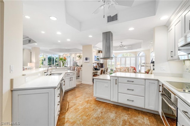 kitchen featuring stainless steel oven, island range hood, sink, a raised ceiling, and kitchen peninsula