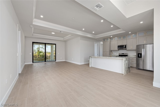 kitchen featuring gray cabinets, a tray ceiling, appliances with stainless steel finishes, a center island with sink, and light stone counters