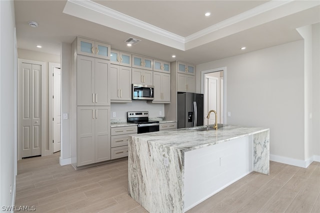 kitchen with light stone countertops, stainless steel appliances, sink, a raised ceiling, and an island with sink