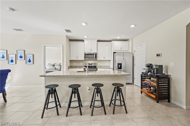 kitchen with an island with sink, stainless steel appliances, light tile floors, white cabinetry, and light stone counters