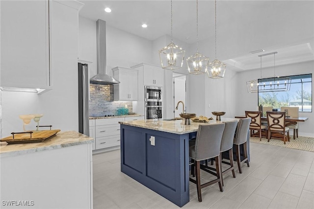 kitchen with white cabinets, a center island with sink, wall chimney range hood, light stone countertops, and decorative light fixtures