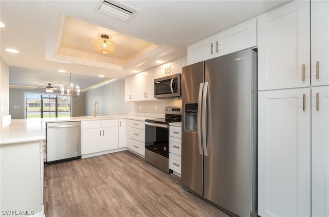 kitchen with white cabinetry, ceiling fan with notable chandelier, appliances with stainless steel finishes, light hardwood / wood-style floors, and a raised ceiling