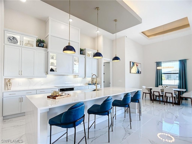 kitchen featuring a breakfast bar, white cabinets, hanging light fixtures, a tray ceiling, and a center island with sink