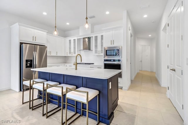 kitchen featuring wall chimney exhaust hood, stainless steel appliances, an island with sink, pendant lighting, and white cabinets