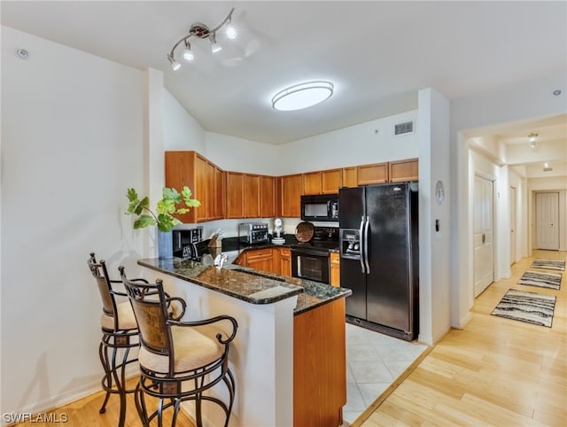 kitchen featuring kitchen peninsula, dark stone countertops, a breakfast bar, light hardwood / wood-style floors, and black appliances