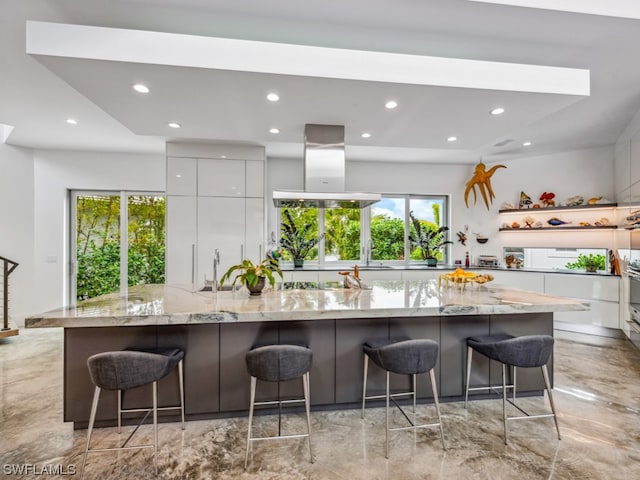 kitchen with a kitchen bar, light stone countertops, island range hood, white cabinets, and a large island