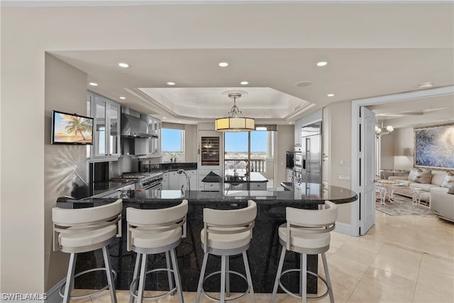 kitchen featuring kitchen peninsula, decorative light fixtures, a tray ceiling, wall chimney exhaust hood, and a breakfast bar area