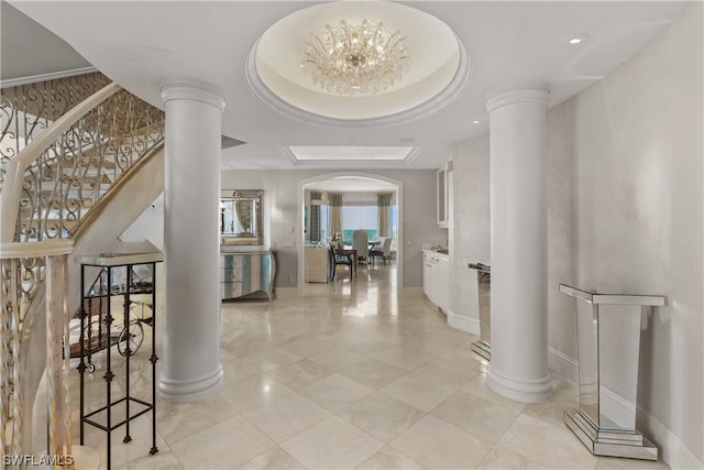 corridor featuring a raised ceiling, an inviting chandelier, decorative columns, and light tile floors