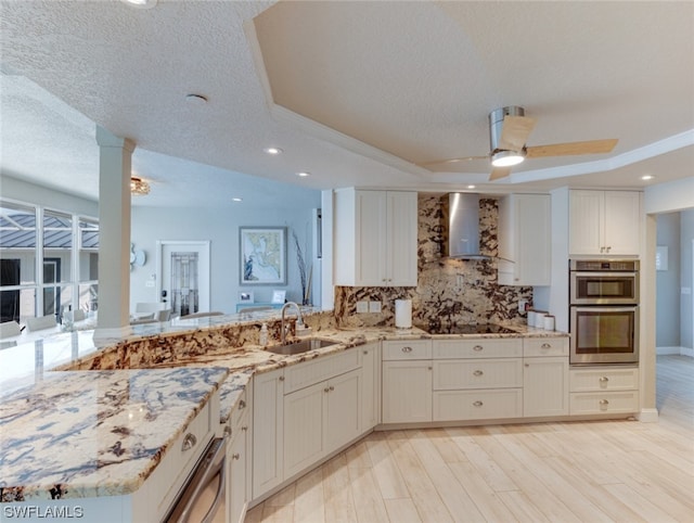 kitchen with a tray ceiling, double oven, ceiling fan, and wall chimney exhaust hood