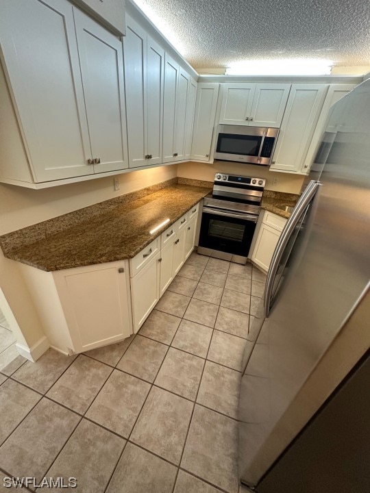 kitchen with dark stone counters, stainless steel appliances, light tile flooring, a textured ceiling, and white cabinetry