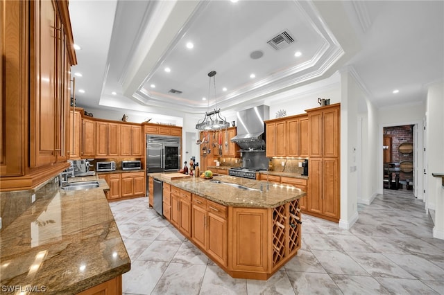 kitchen with an island with sink, sink, light stone counters, a tray ceiling, and wall chimney exhaust hood