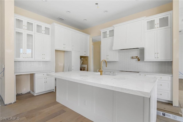 kitchen with decorative backsplash, light stone counters, light hardwood / wood-style flooring, white cabinetry, and an island with sink