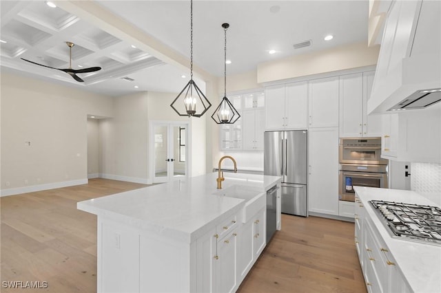 kitchen featuring stainless steel appliances, white cabinets, hanging light fixtures, an island with sink, and custom range hood