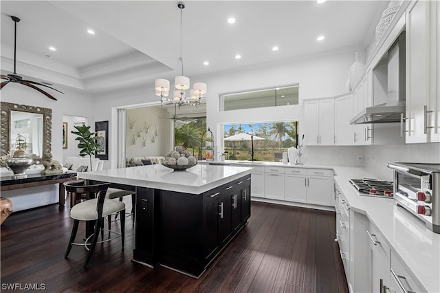 kitchen featuring pendant lighting, a center island, ceiling fan with notable chandelier, wall chimney exhaust hood, and dark hardwood / wood-style flooring