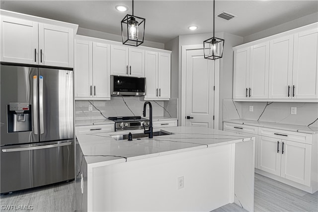 kitchen featuring appliances with stainless steel finishes, light stone counters, white cabinets, an island with sink, and decorative light fixtures