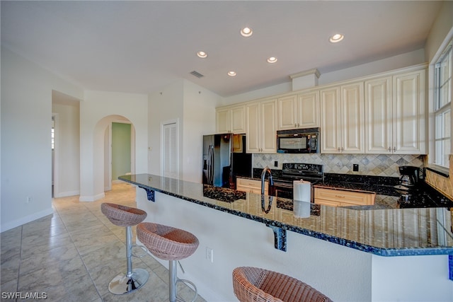 kitchen with light tile floors, a breakfast bar, black appliances, backsplash, and dark stone countertops