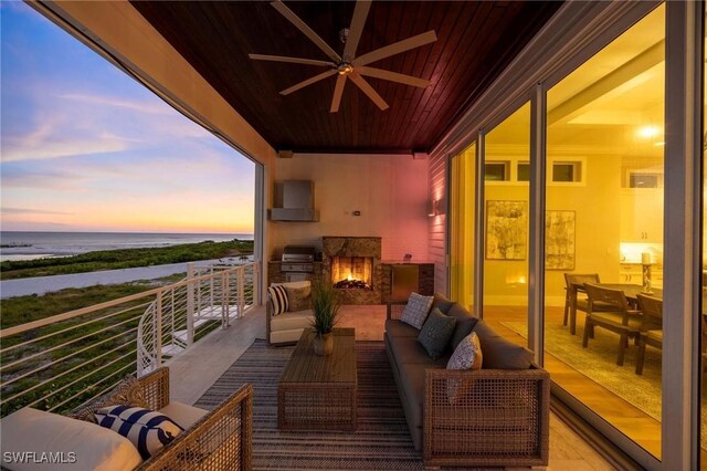 balcony at dusk featuring an outdoor living space with a fireplace, a grill, ceiling fan, and a water view