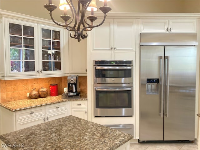 kitchen with backsplash, dark stone counters, stainless steel appliances, an inviting chandelier, and hanging light fixtures