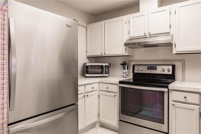 kitchen with under cabinet range hood, stainless steel appliances, white cabinetry, tile counters, and decorative backsplash