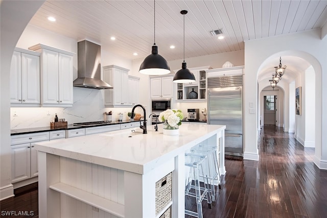 kitchen featuring wall chimney exhaust hood, built in appliances, white cabinets, decorative light fixtures, and a large island with sink