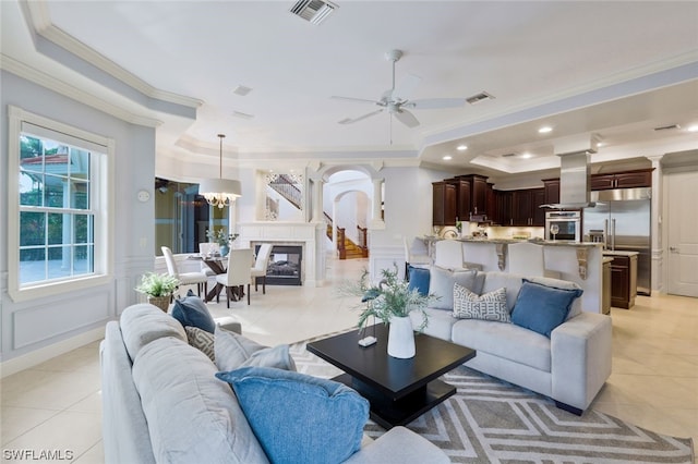 living room with crown molding, a raised ceiling, light tile floors, and ceiling fan with notable chandelier