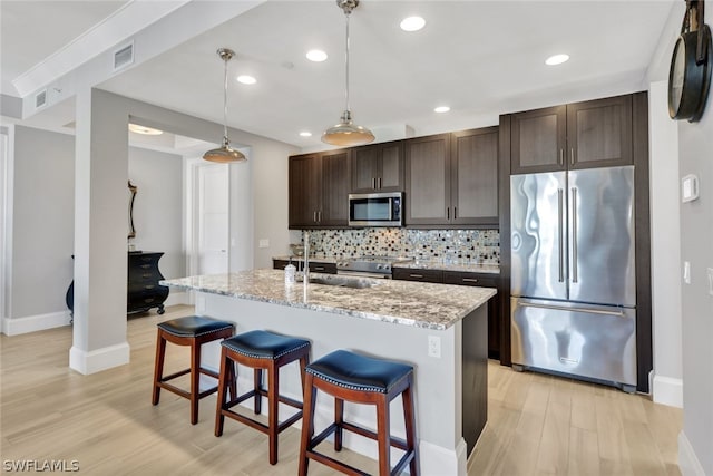 kitchen featuring appliances with stainless steel finishes, a center island with sink, light stone counters, decorative light fixtures, and sink