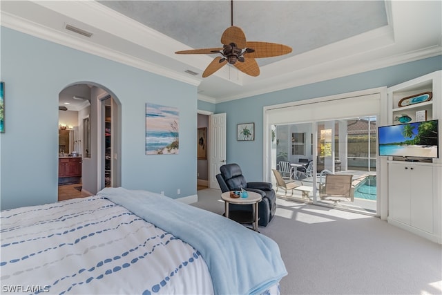 bedroom featuring light colored carpet, ornamental molding, a tray ceiling, ensuite bath, and ceiling fan with notable chandelier