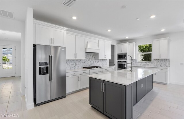 kitchen with custom range hood, stainless steel appliances, white cabinetry, and a kitchen island with sink