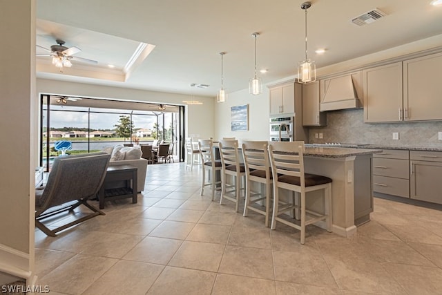 kitchen with pendant lighting, premium range hood, a kitchen bar, ceiling fan, and a tray ceiling