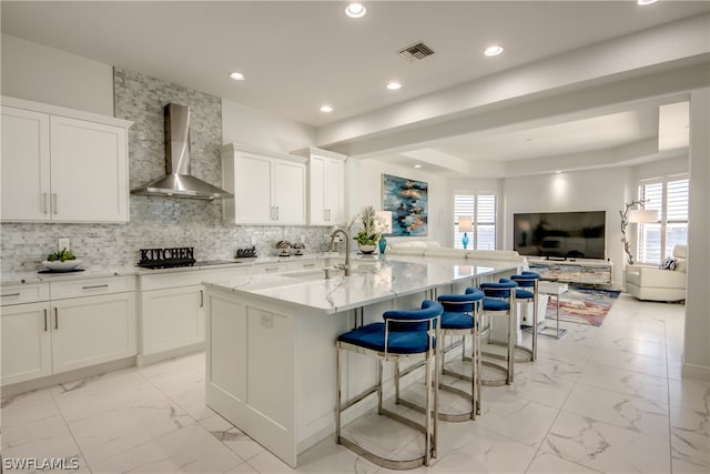 kitchen featuring an island with sink, wall chimney range hood, a breakfast bar area, white cabinets, and sink