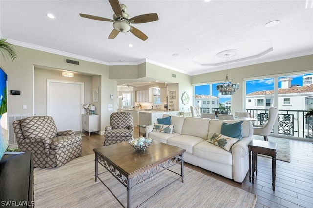 living room with a raised ceiling, crown molding, light hardwood / wood-style flooring, and ceiling fan with notable chandelier