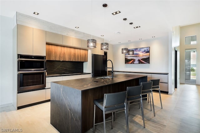 kitchen featuring sink, double oven, a center island with sink, decorative light fixtures, and a kitchen bar