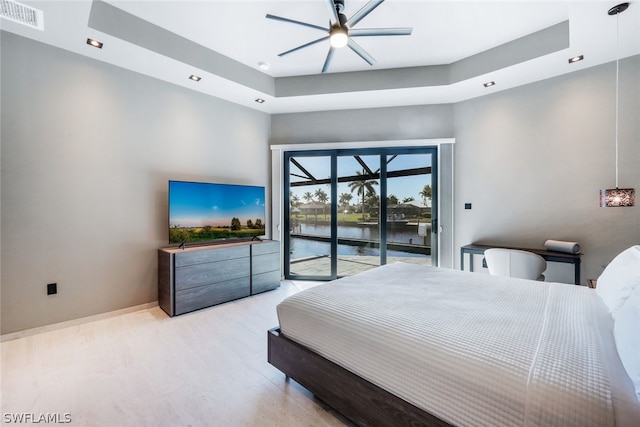 bedroom with a tray ceiling, ceiling fan, access to exterior, light hardwood / wood-style floors, and a water view