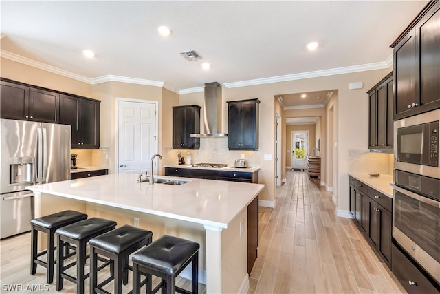 kitchen with light hardwood / wood-style flooring, backsplash, appliances with stainless steel finishes, wall chimney range hood, and a center island with sink