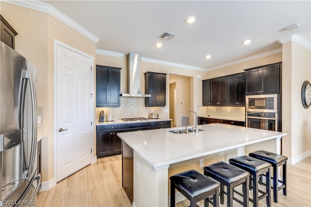 kitchen with wall chimney exhaust hood, light wood-type flooring, stainless steel appliances, a center island with sink, and sink