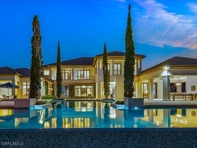 back house at dusk featuring a patio, a balcony, and a pool with hot tub