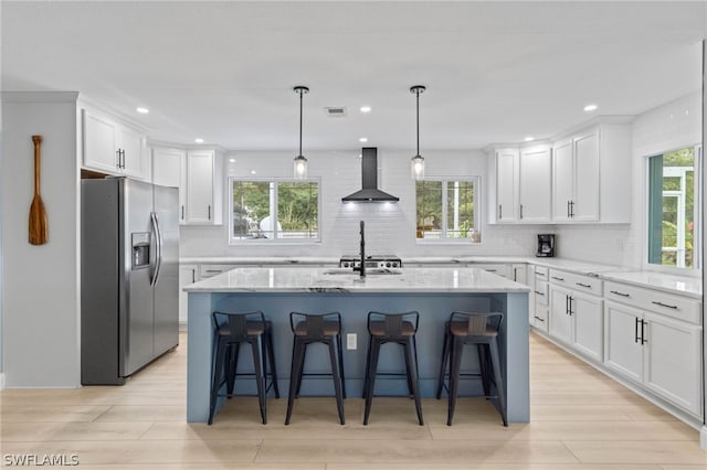 kitchen featuring stainless steel refrigerator with ice dispenser, wall chimney range hood, an island with sink, and a kitchen bar