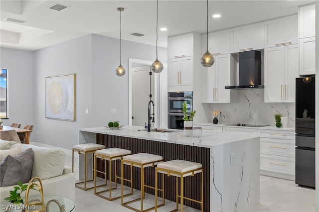 kitchen with light stone counters, a center island with sink, wall chimney exhaust hood, sink, and stainless steel double oven