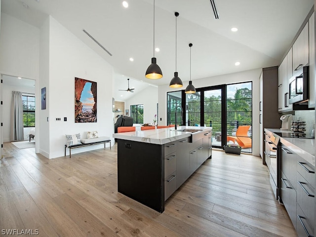 kitchen with an island with sink, ceiling fan, hanging light fixtures, stainless steel microwave, and light hardwood / wood-style floors